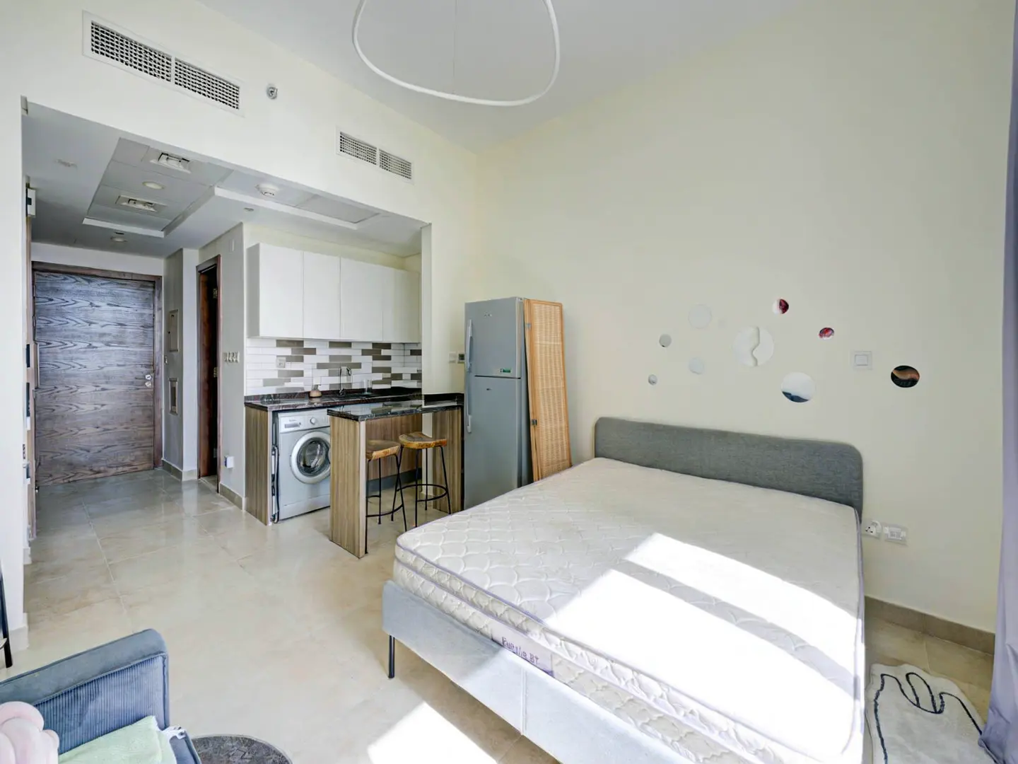 Studio apartment with a bed, kitchen, and washing machine. The walls are cream-colored, and the floor is tiled.