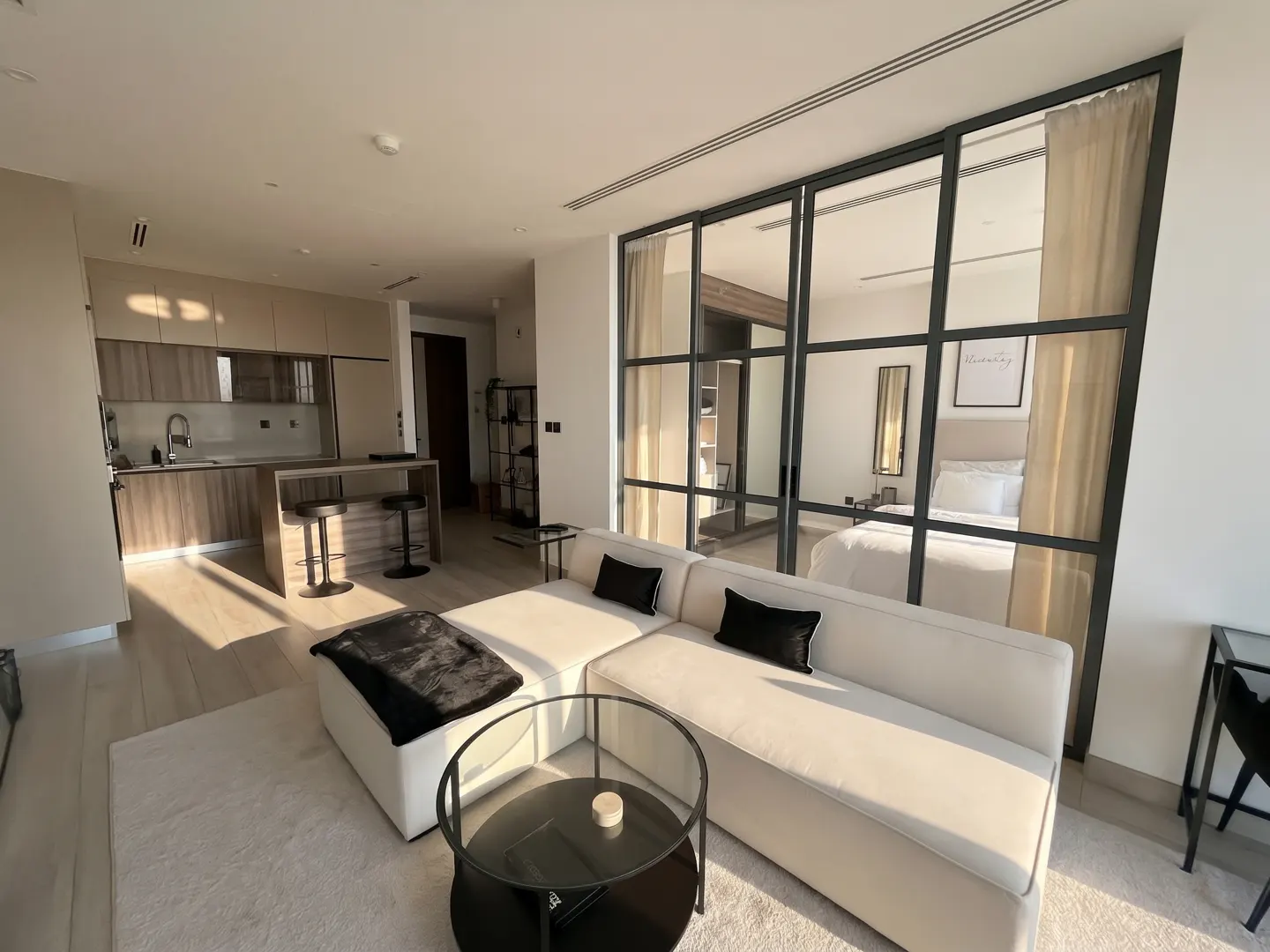 Bright, modern apartment interior with a white sectional sofa, black pillows, and a glass coffee table. A kitchen and bedroom are visible in the background.