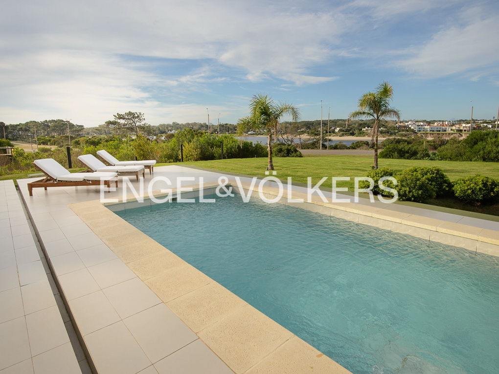 A rectangular pool with turquoise water, surrounded by a tiled patio, lawn, and palm trees under a blue sky. Three white lounge chairs are on the patio.