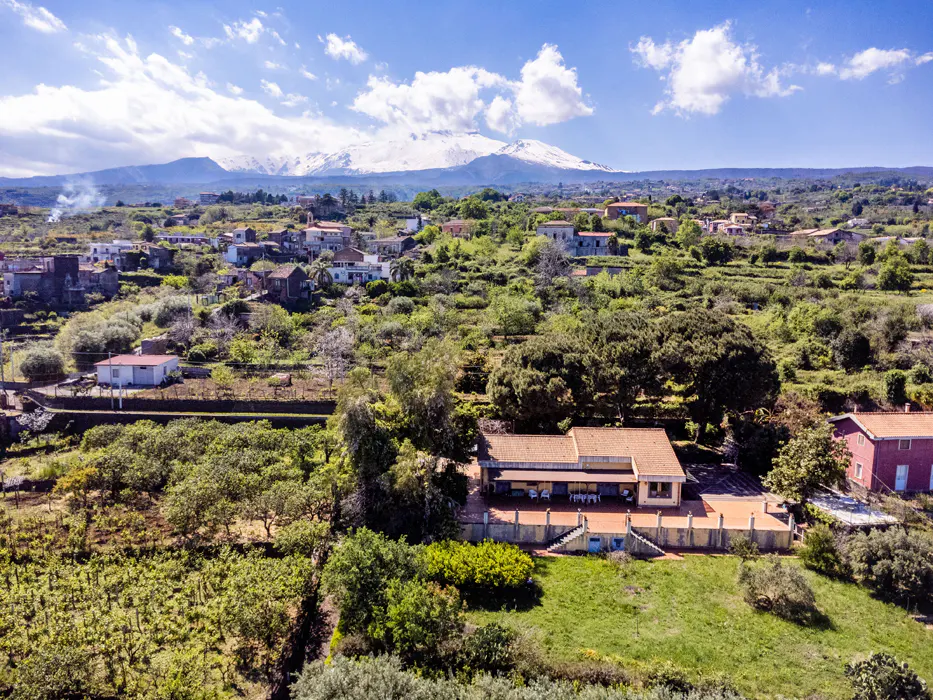 Scenic view of a property with a red tile roof, surrounded by lush greenery and trees. In the background, a snow-capped mountain under a blue sky.