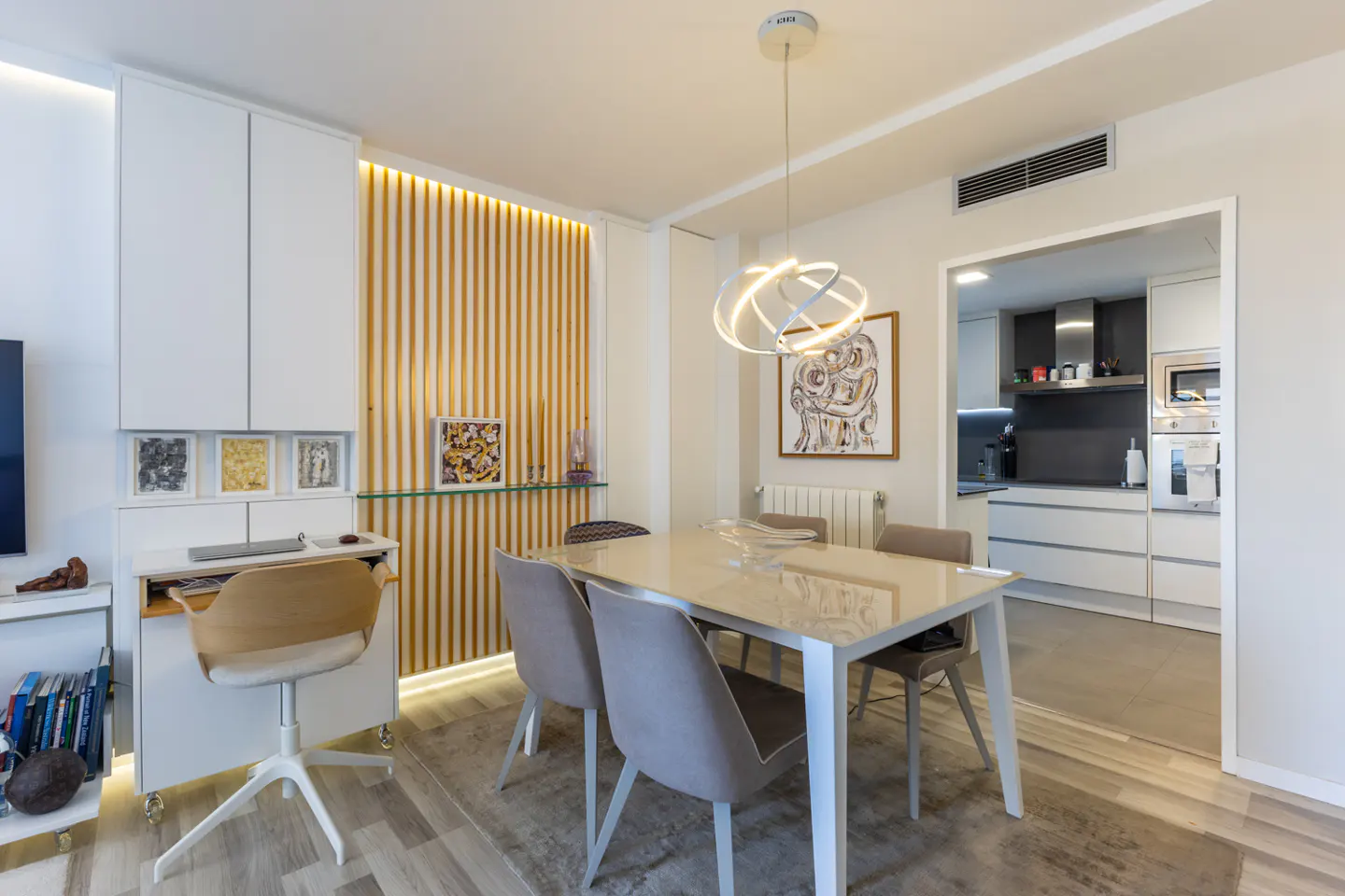 Modern dining area with a glass table, four grey chairs, a unique pendant light, and a view into a white kitchen.