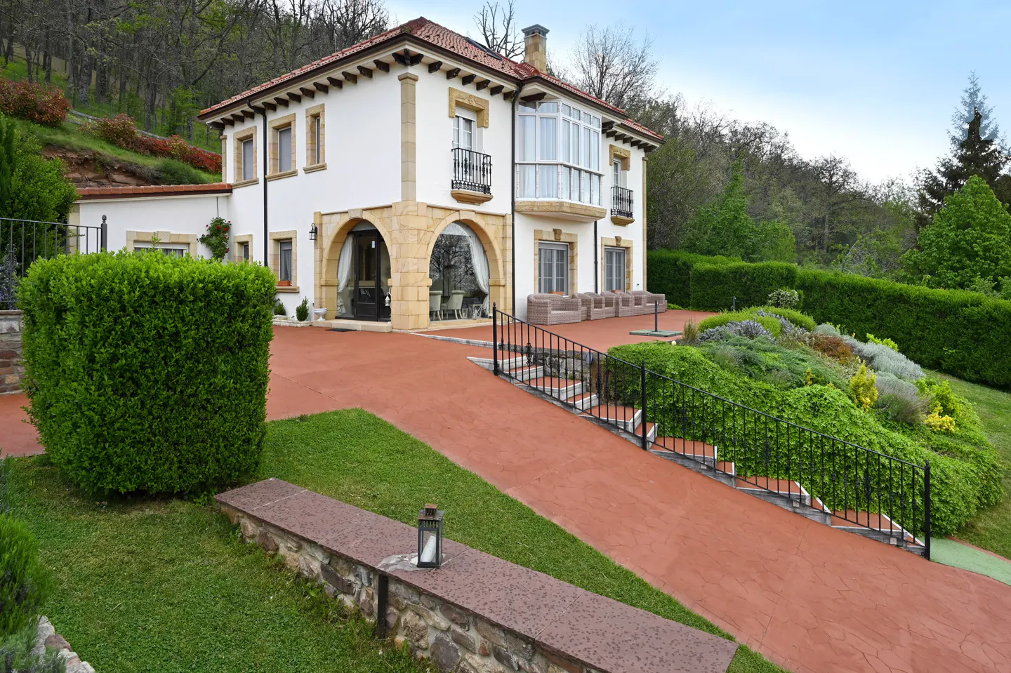 A two-story white house with a red tile roof and a red driveway surrounded by green landscaping.