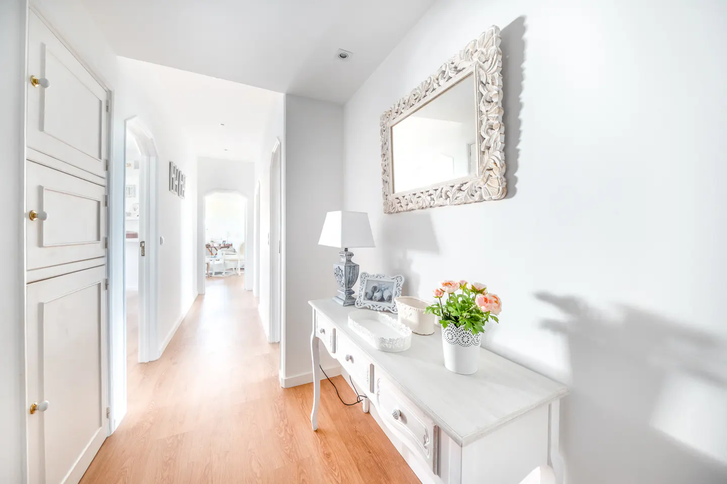 Bright hallway with wood floors, white walls, and a white console table. A decorative mirror hangs above the table, which holds a lamp and flowers.