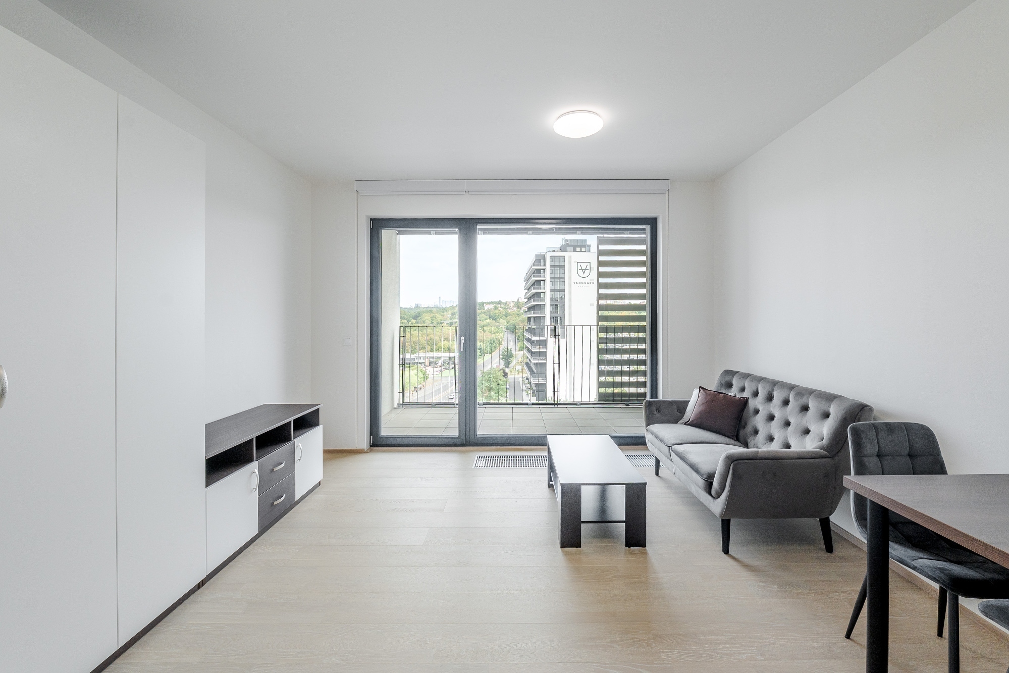 Modern living room with light wood floors, a grey tufted sofa, coffee table, and balcony access with city views. [AI-generated text]