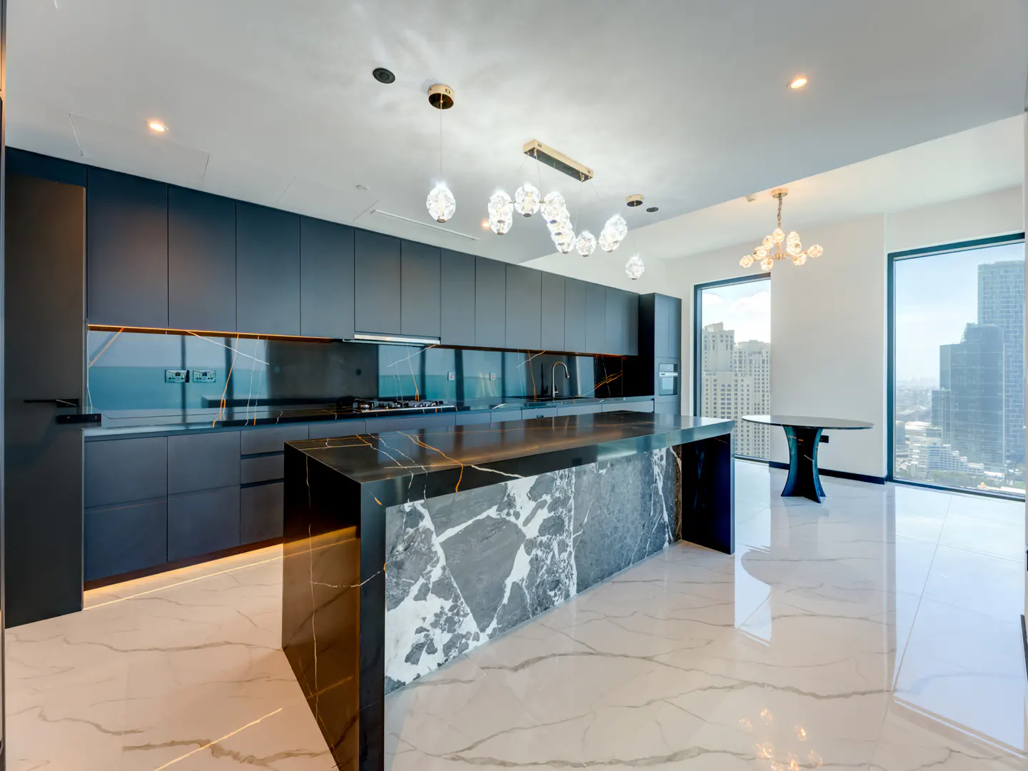 A modern kitchen with dark cabinets, marble island, and city view. Crystal chandeliers hang above.