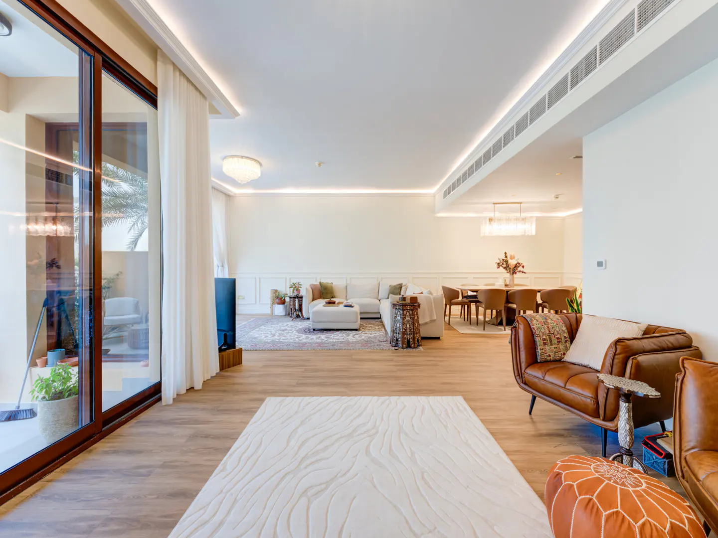Bright, modern living room with light wood floors, white walls, and a large glass door to a patio. Furnished with brown leather chairs, a white sofa, and a dining table.
