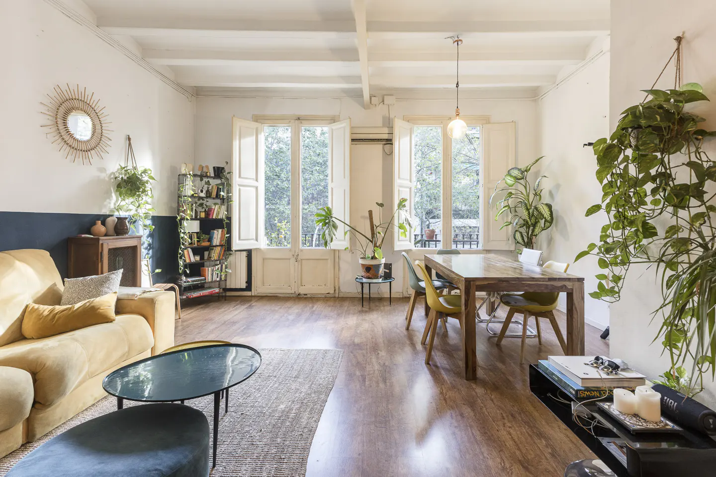 Bright living room with a yellow sofa, wooden table, and lots of plants. White walls and wooden floors create a cozy atmosphere.