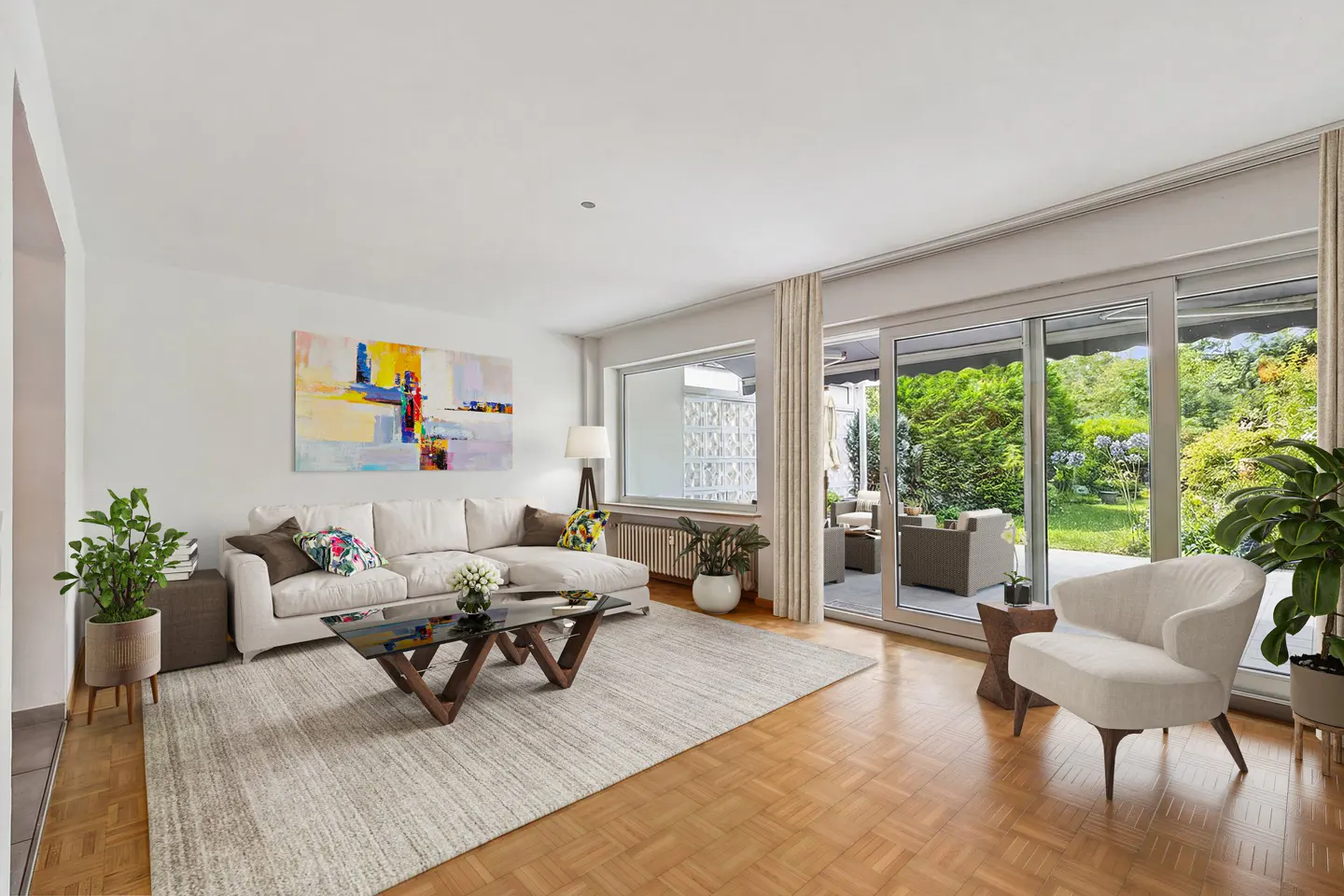Bright living room with white sofa, armchair, and glass table on a light rug. Sliding glass doors open to a patio and garden. Abstract art on the wall.