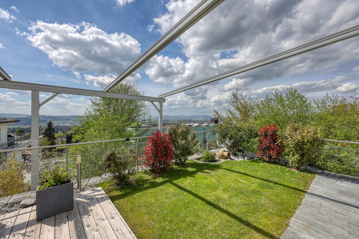 A backyard with green grass, red bushes, and a wooden deck under a cloudy sky.