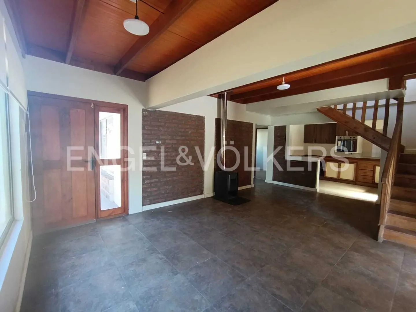 Open-plan living space with wood ceiling, brick accent wall, and wood-burning stove. Stairs lead to an upper level. Kitchen in background.
