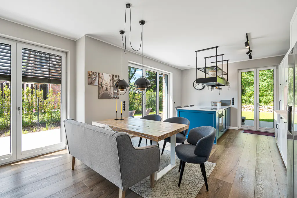 A modern kitchen and dining area with a wooden table, gray chairs, and a blue kitchen island. Large windows offer views of the garden.