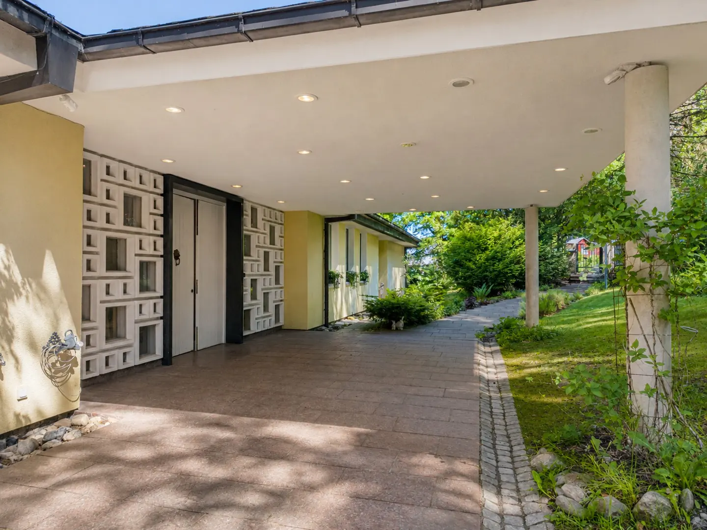 Covered carport of a modern home with yellow walls, patterned concrete blocks, and a stone walkway leading to a green lawn.