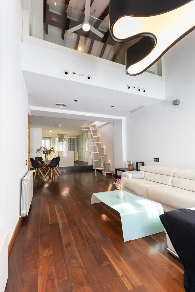 Bright, modern living room with hardwood floors, white walls, and a white sofa. A glass coffee table sits in front of the sofa. Stairs lead to a loft area.