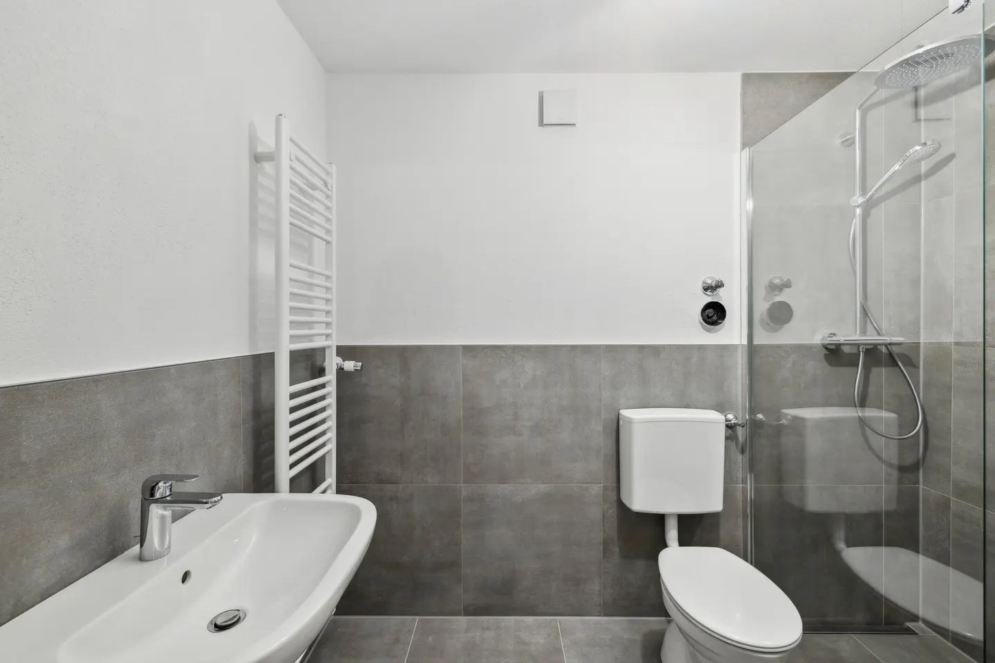 Bathroom with white sink, toilet, and towel rack. Gray tile wainscoting and glass shower enclosure.