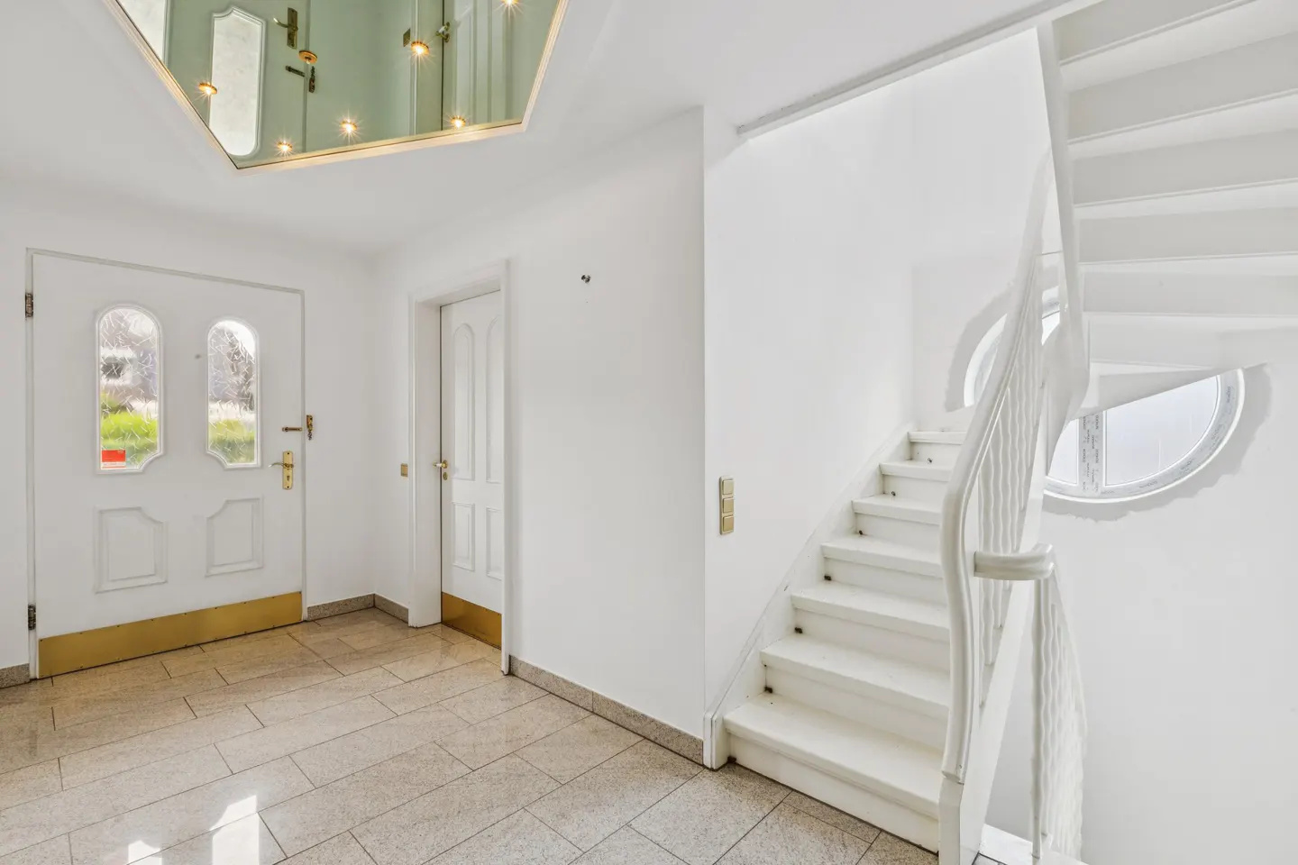Bright foyer with white walls, tiled floor, and a white staircase with a curved railing. A front door with glass panels is visible.