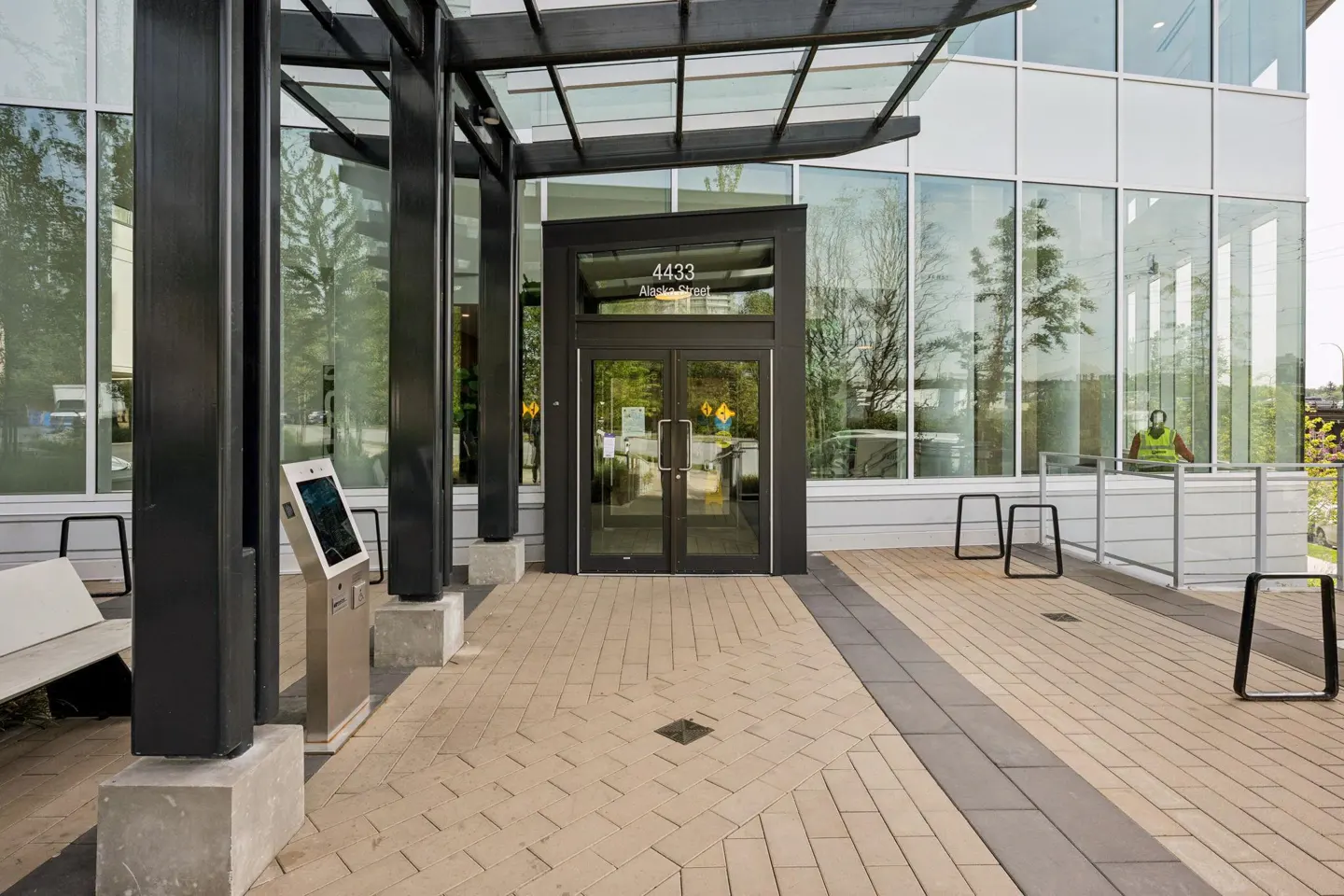 Exterior of a modern building with glass windows and a black framed entrance. The address "4433 Alaska Street" is above the doors. A kiosk and bench are to the left.