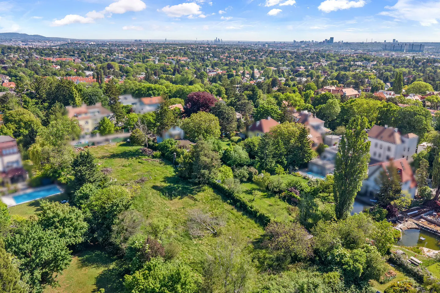 Aerial view of a lush green property with trees, grass, and houses, under a blue sky with scattered clouds. A city skyline is visible in the distance.