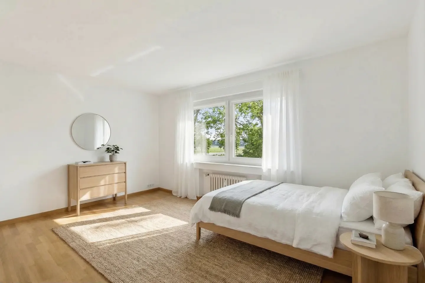 Bright bedroom with white walls, wood floor, and a jute rug. A bed with white linens sits near a window with sheer curtains. A dresser with a round mirror is on the left.