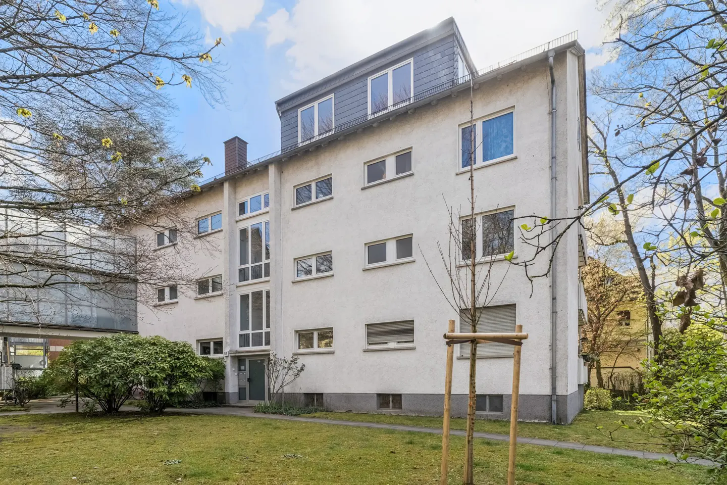 Exterior view of a three-story white apartment building with a gray roof, surrounded by green lawn and trees.