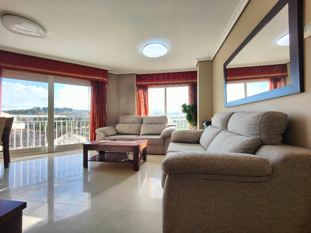 Bright living room with beige sofas, a wooden coffee table, tile floors, and large windows with red curtains.