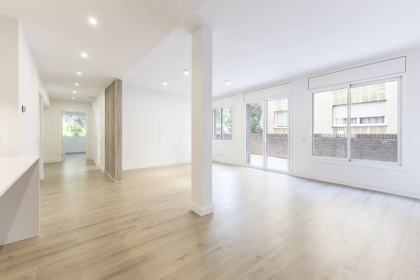 Bright, open-plan living area with light wood floors, white walls, a central pillar, and large windows overlooking a patio.
