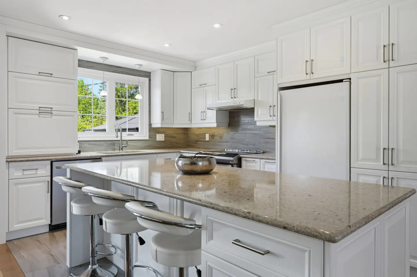 Bright kitchen with white cabinets, stainless steel appliances, and a large island with white stools. A window overlooks a green yard.