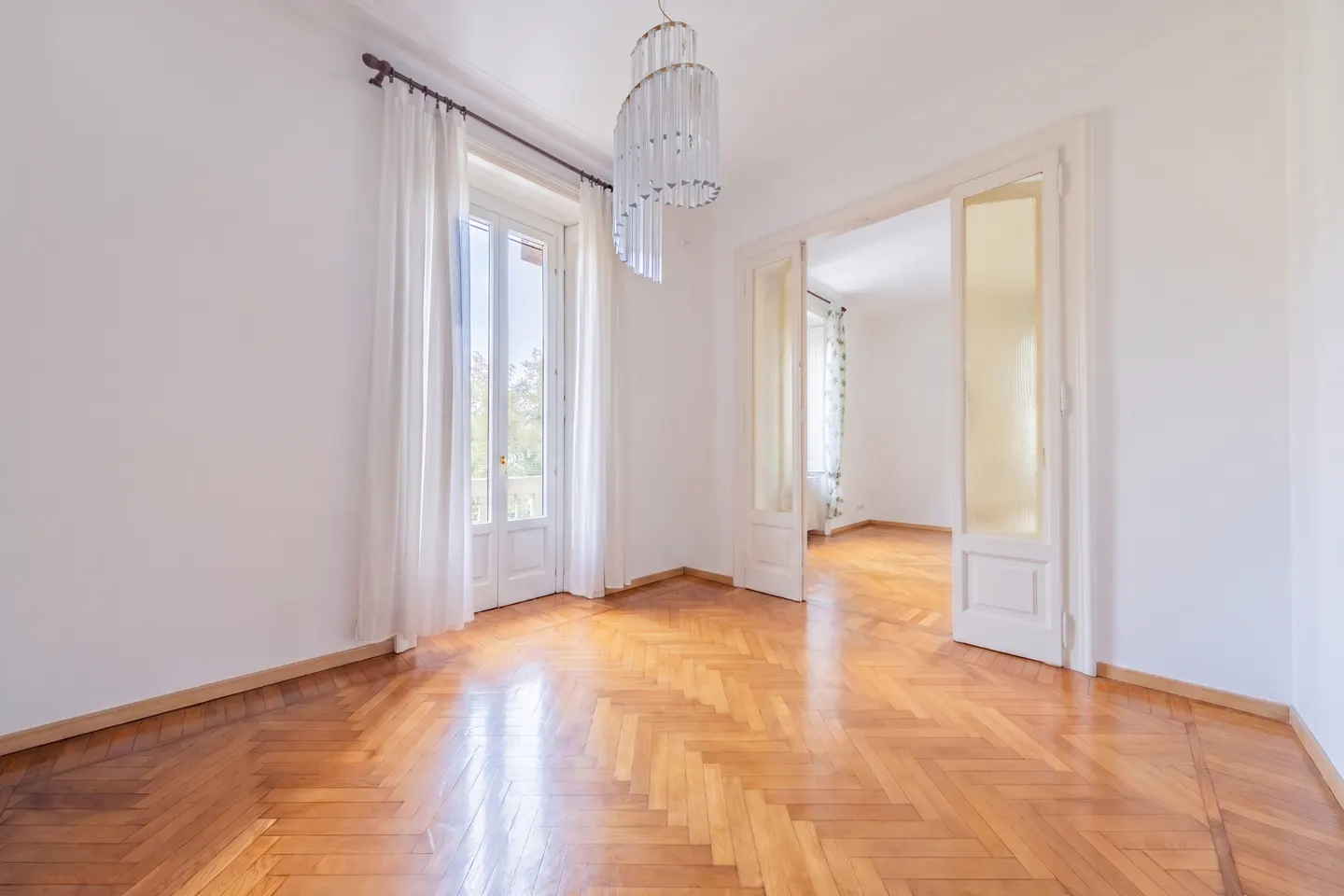 Bright, empty room with herringbone wood floors, white walls, and a glass chandelier. A window with white curtains and an open doorway let in natural light.