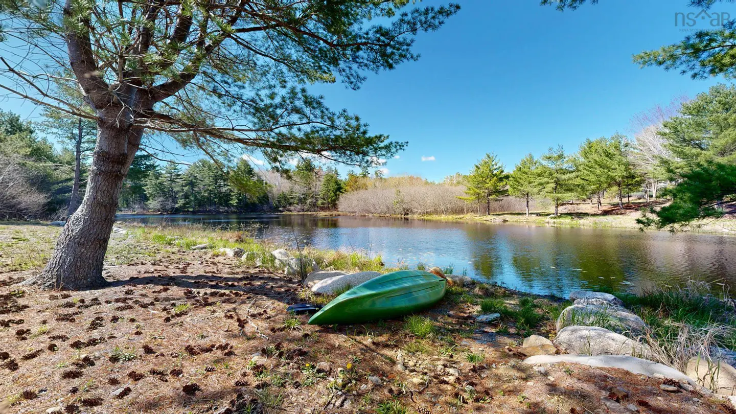 A green kayak sits on the shore of a calm lake under a clear blue sky. Pine trees surround the water, and pine cones litter the ground.