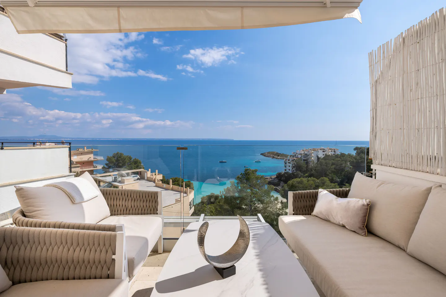 Balcony with beige sofas, white table, and ocean view. Blue sky and turquoise water visible beyond the glass railing.