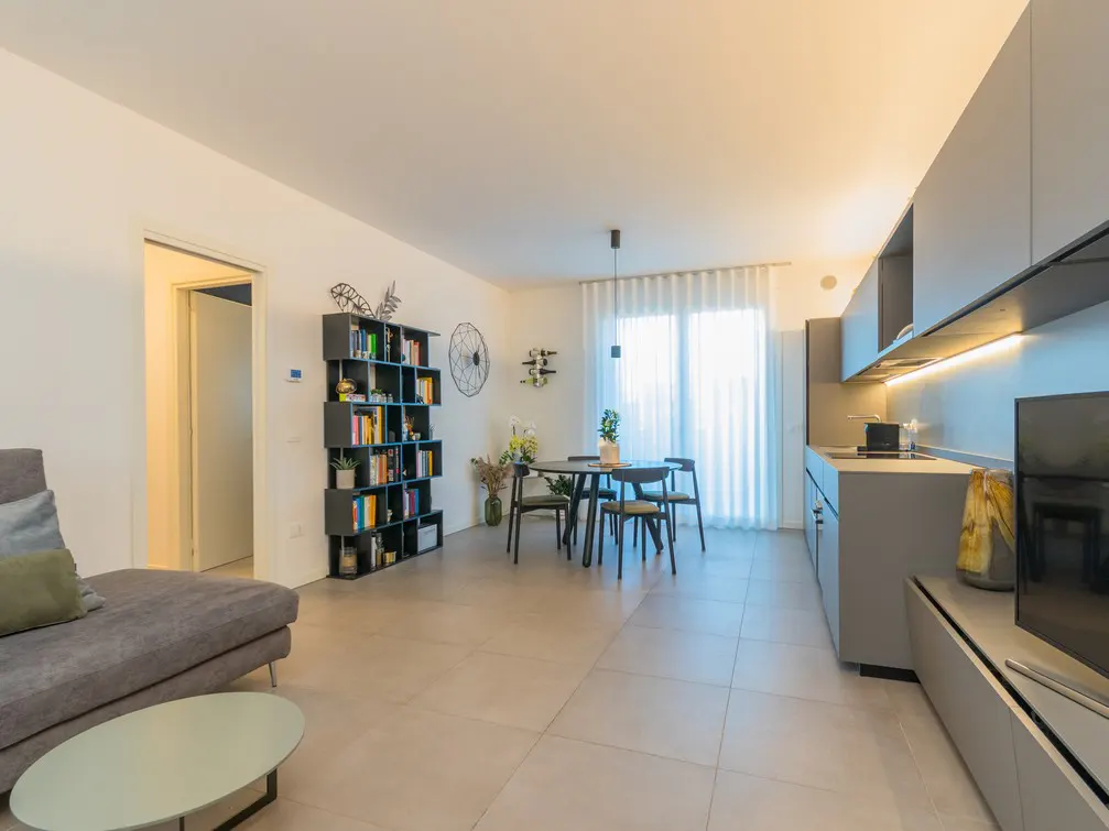 Modern open-plan living area with a grey sofa, dining table, bookshelf, and sleek grey kitchen cabinets.