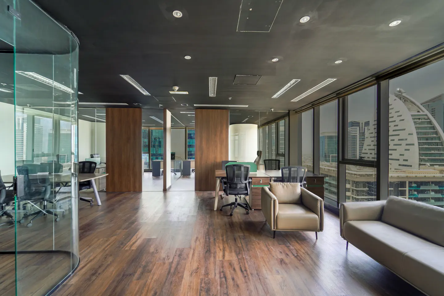 Office space with wood floors, glass walls, desks, and chairs. A tan sofa and chair sit near large windows with a city view.