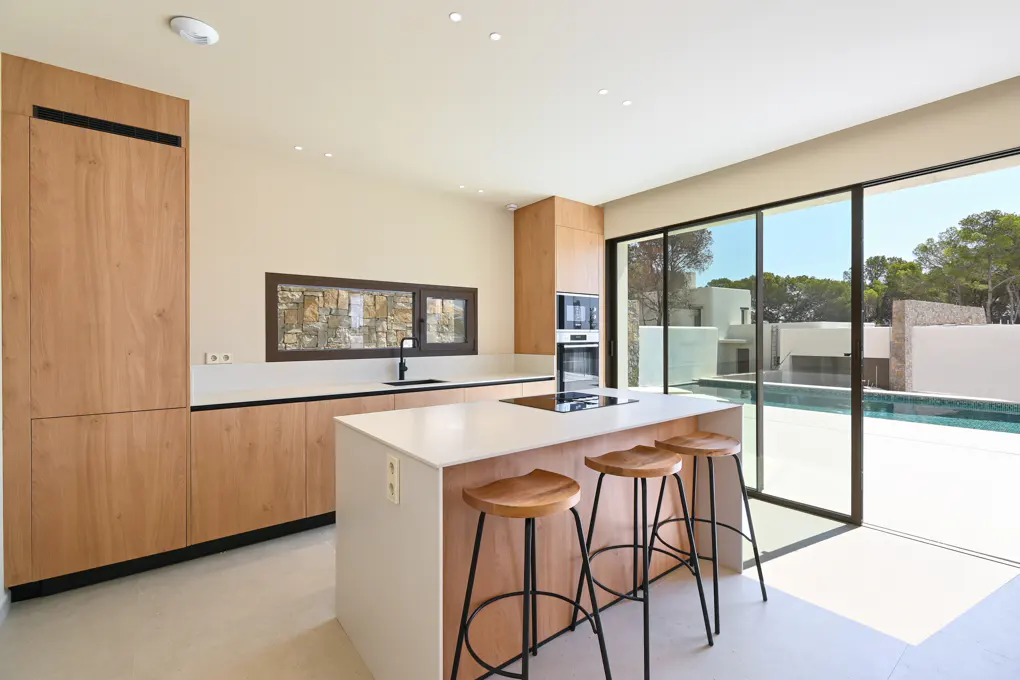 A modern kitchen with wood cabinets, a white island with three stools, and a view of a pool through sliding glass doors.