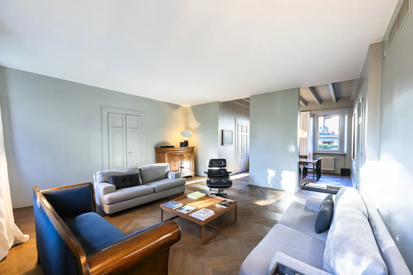 Living room with gray walls, herringbone wood floors, sofas, a blue armchair, and a wooden coffee table with books.