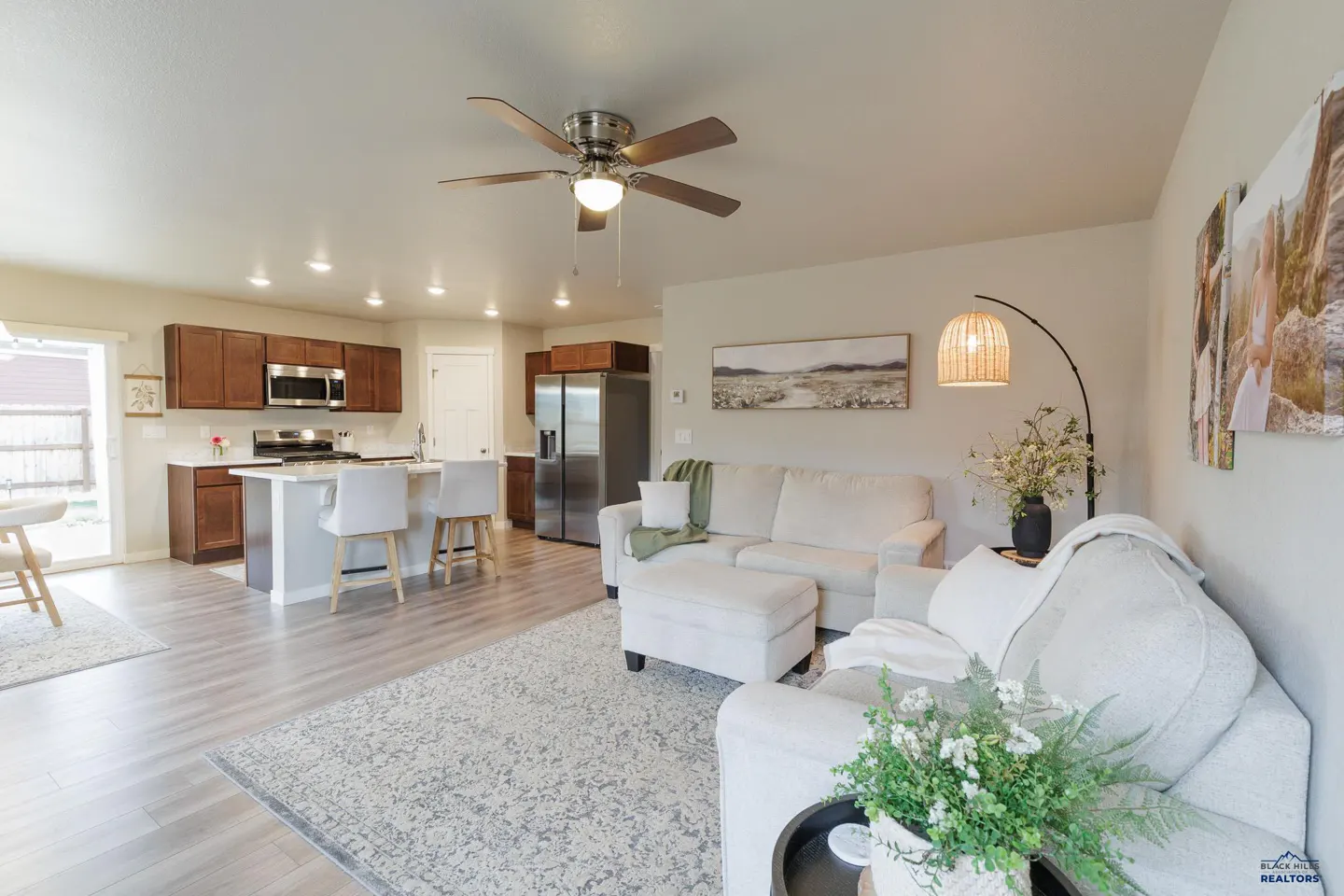 Open-concept living space with a kitchen, white sofa, and a ceiling fan. The kitchen has brown cabinets and stainless steel appliances.
