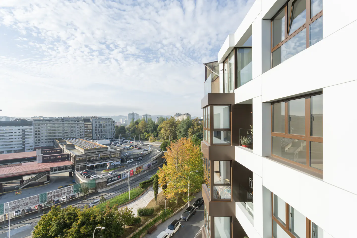 View from a modern white apartment building with brown window frames, overlooking a city with a bus station and autumn trees.