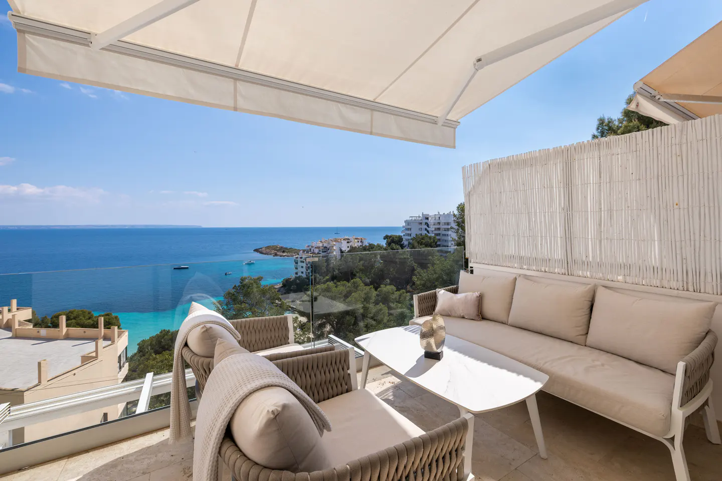 Balcony view with beige furniture, white table, and ocean backdrop. Awnings provide shade. Turquoise water and blue sky visible.