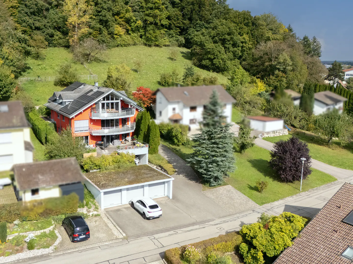 Aerial view of a bright orange house with balconies, solar panels, and a triple garage, set against a lush, wooded hill.