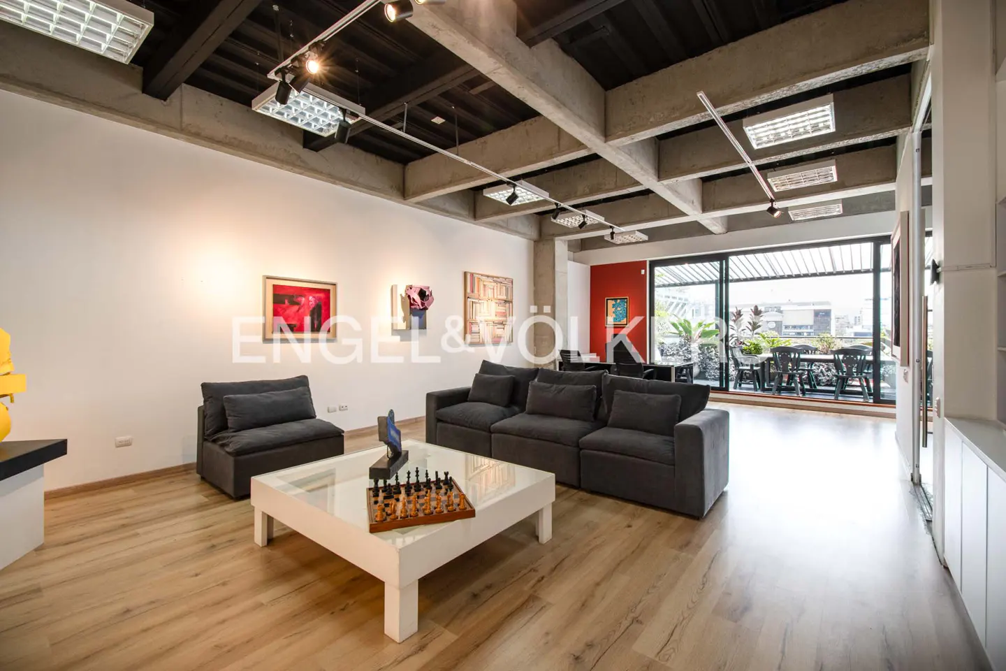 Living room with gray sofa, chair, and white coffee table with chess set. Art on walls, balcony with table and chairs.
