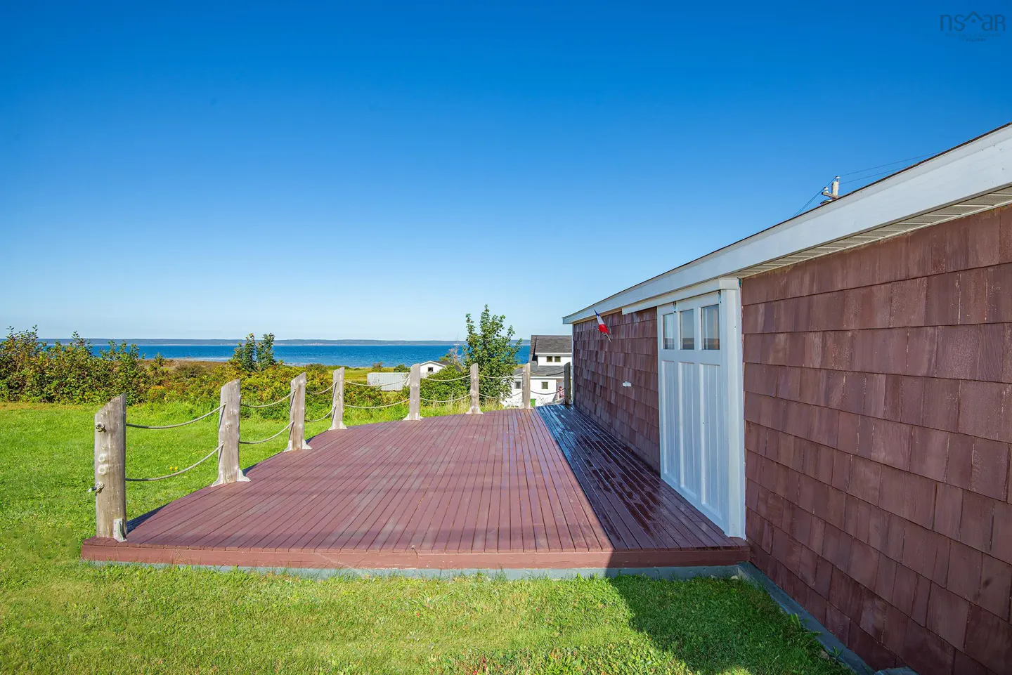 Wooden deck with rope railing overlooks the ocean. The house has brown siding and a white door. Blue sky.