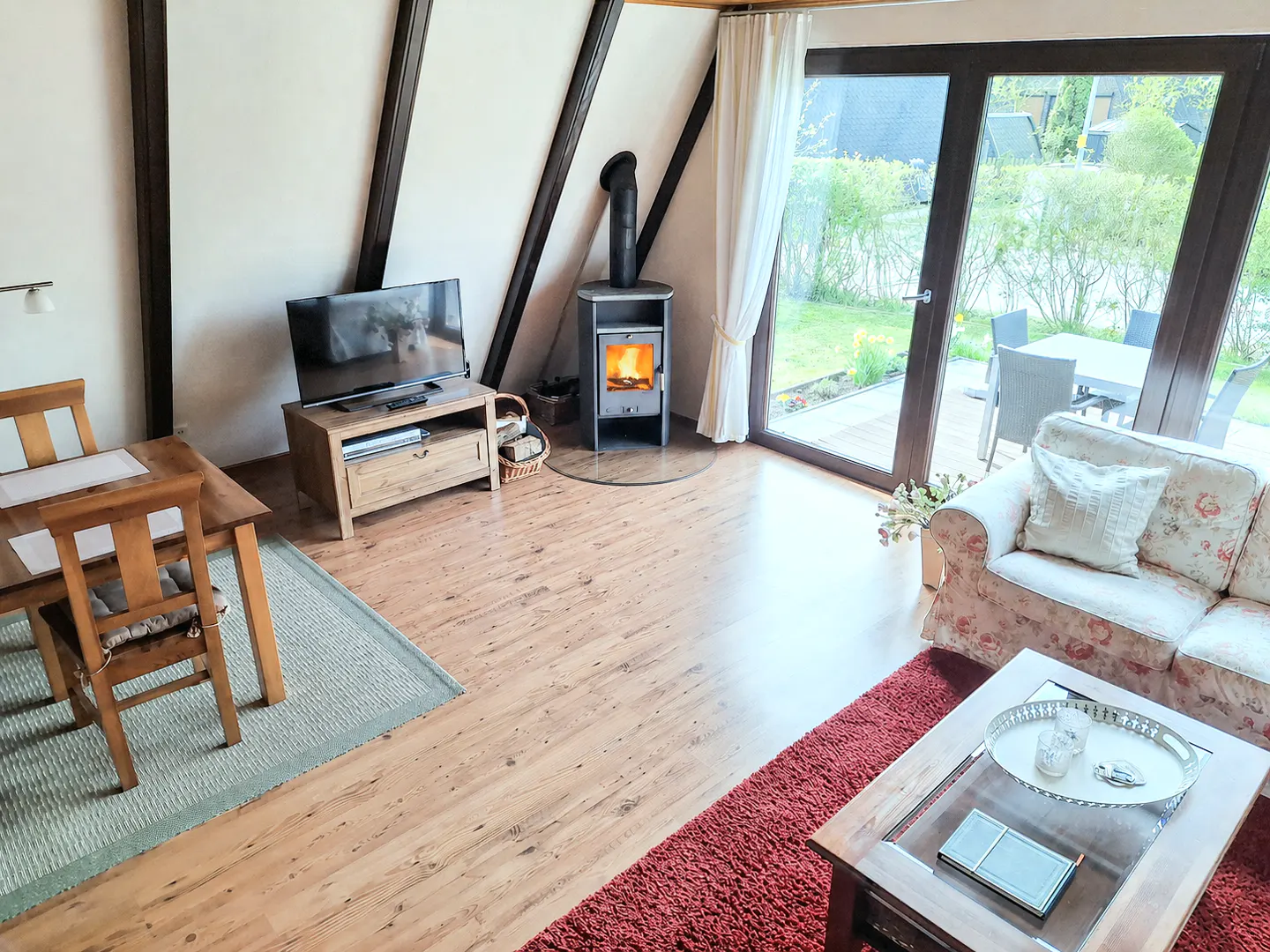Living room with wood floors, a floral sofa, a wood-burning stove, and a dining table. Outside is a patio with a table and chairs.
