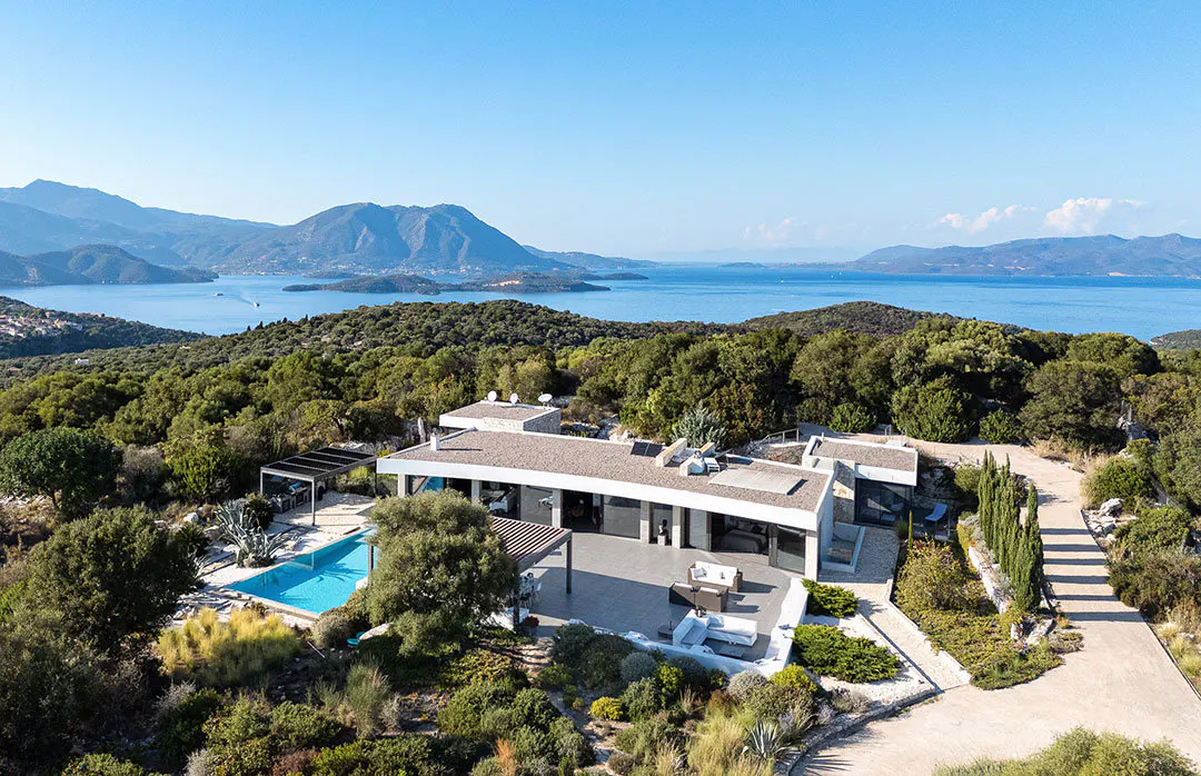 Aerial view of a modern white house with a pool, surrounded by green trees, overlooking the blue ocean and mountains under a clear blue sky.