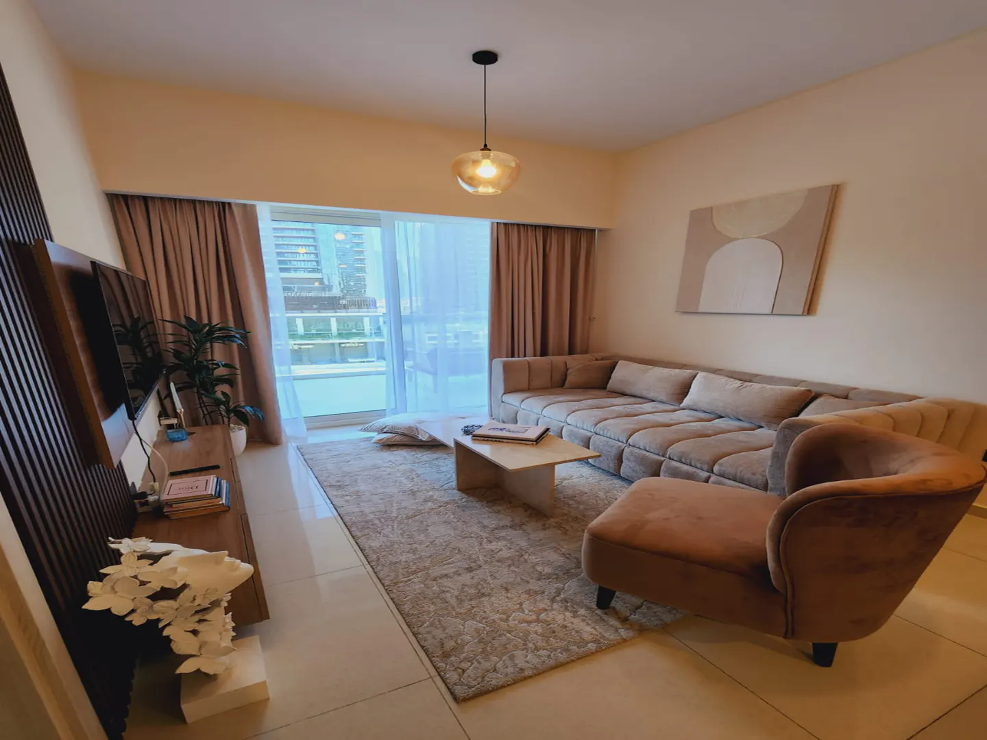 A modern living room with a beige sectional sofa, chaise lounge, and a light-colored rug. A coffee table sits in the center, and a large window offers a city view.