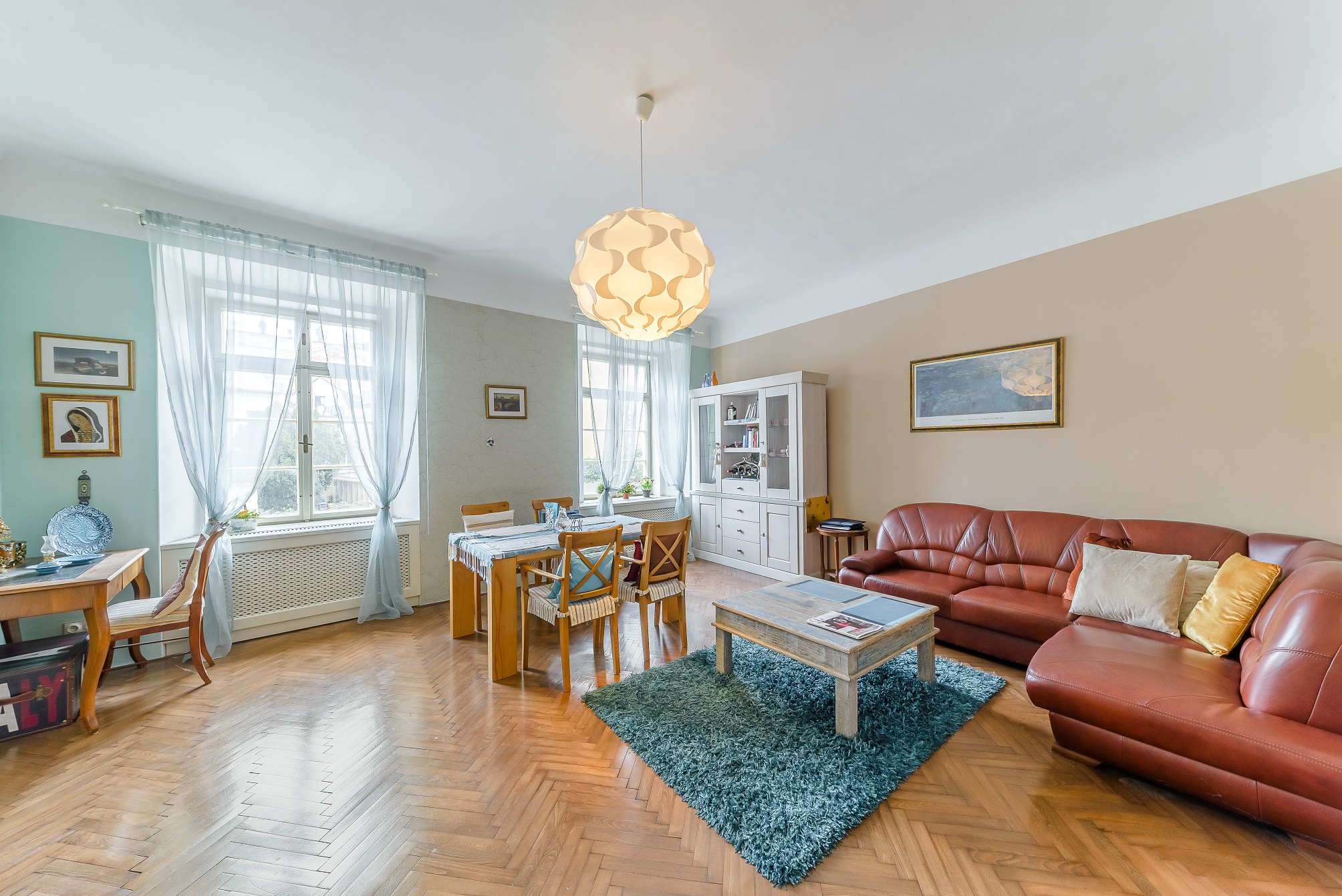 Living room with herringbone wood floors, a brown leather sectional, a blue rug, a dining set, and a white hutch.