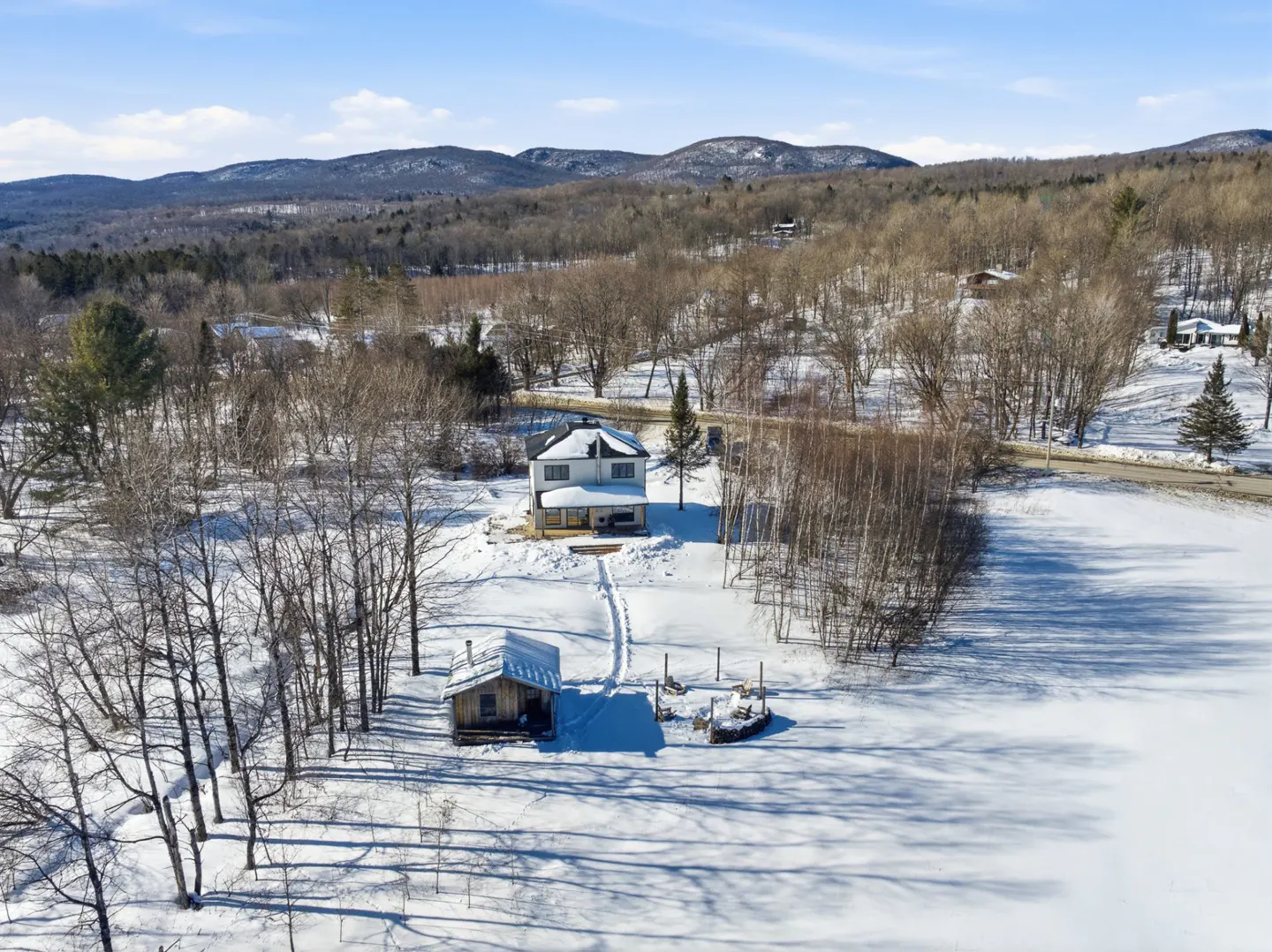 Winter aerial view of a white house with a small cabin on a snow-covered landscape with mountains in the background.