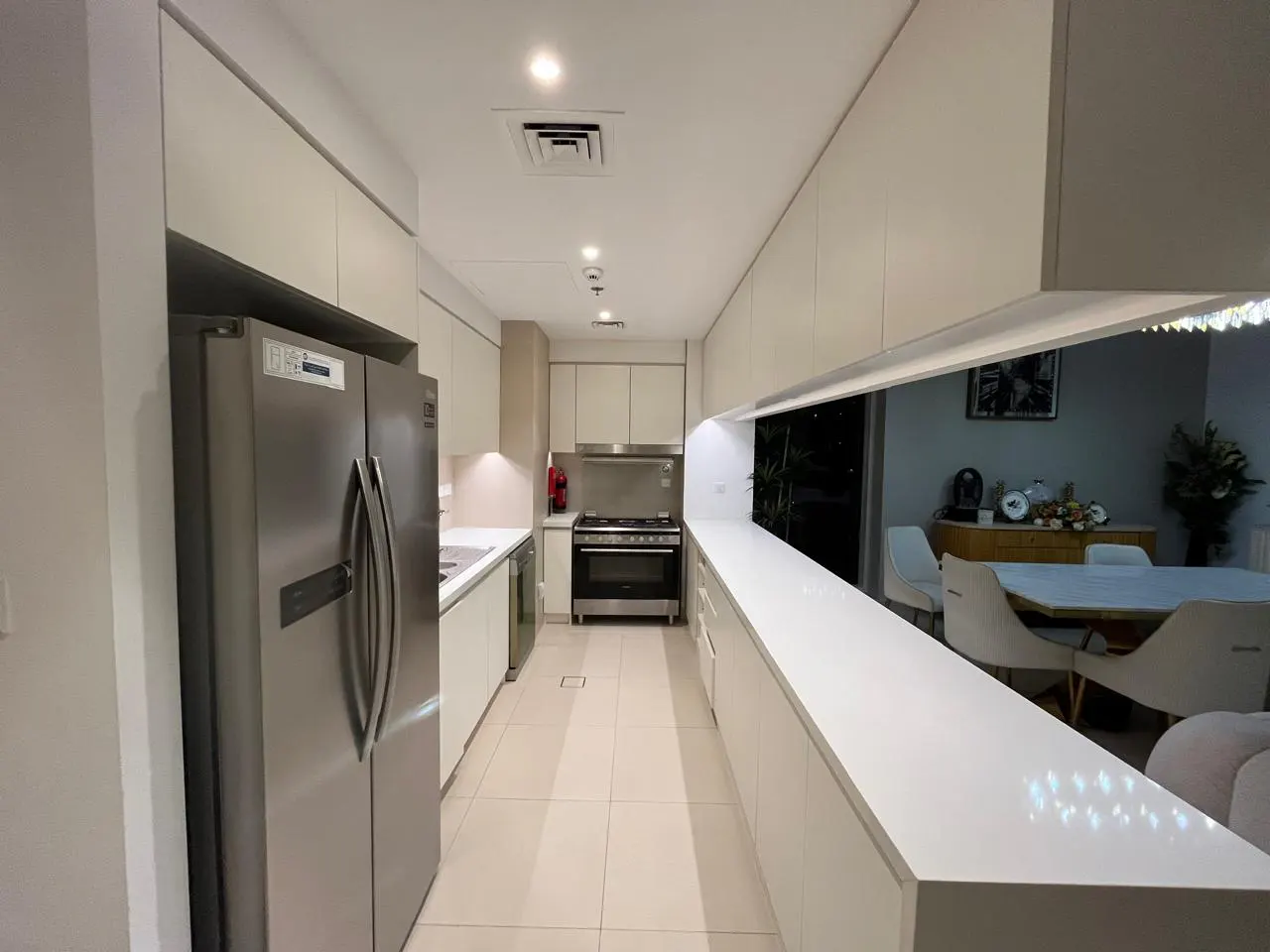 A modern, narrow kitchen with white cabinets, stainless steel appliances, and a pass-through to the dining area.