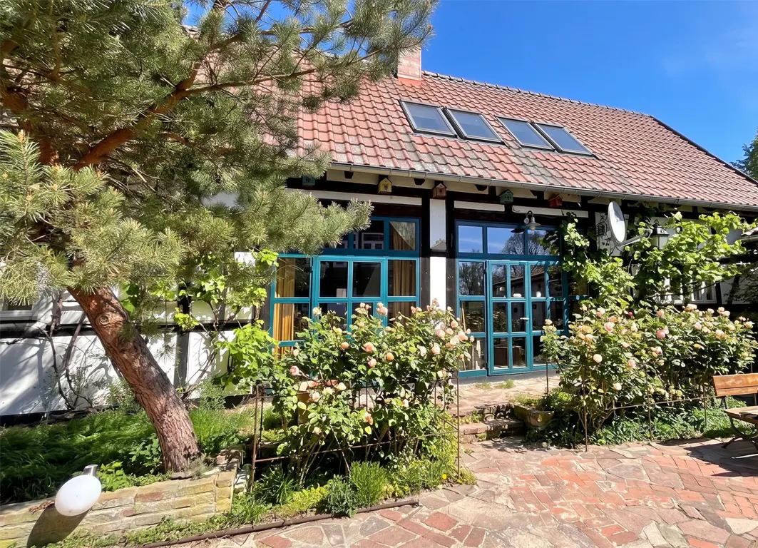 Exterior view of a half-timbered house with a red tiled roof and turquoise framed windows, surrounded by roses and greenery.