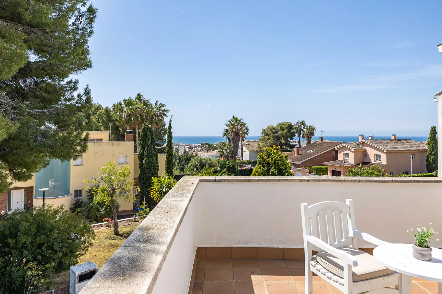 Balcony view with white chair and table, overlooking houses, trees, and the ocean under a clear blue sky.