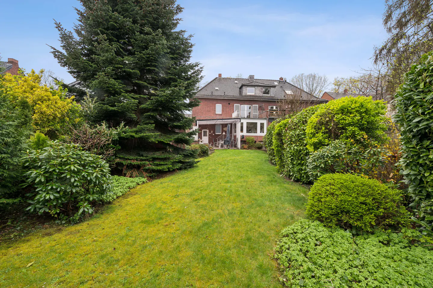 A lush green lawn leads to a red brick house with a balcony, framed by trees and hedges.
