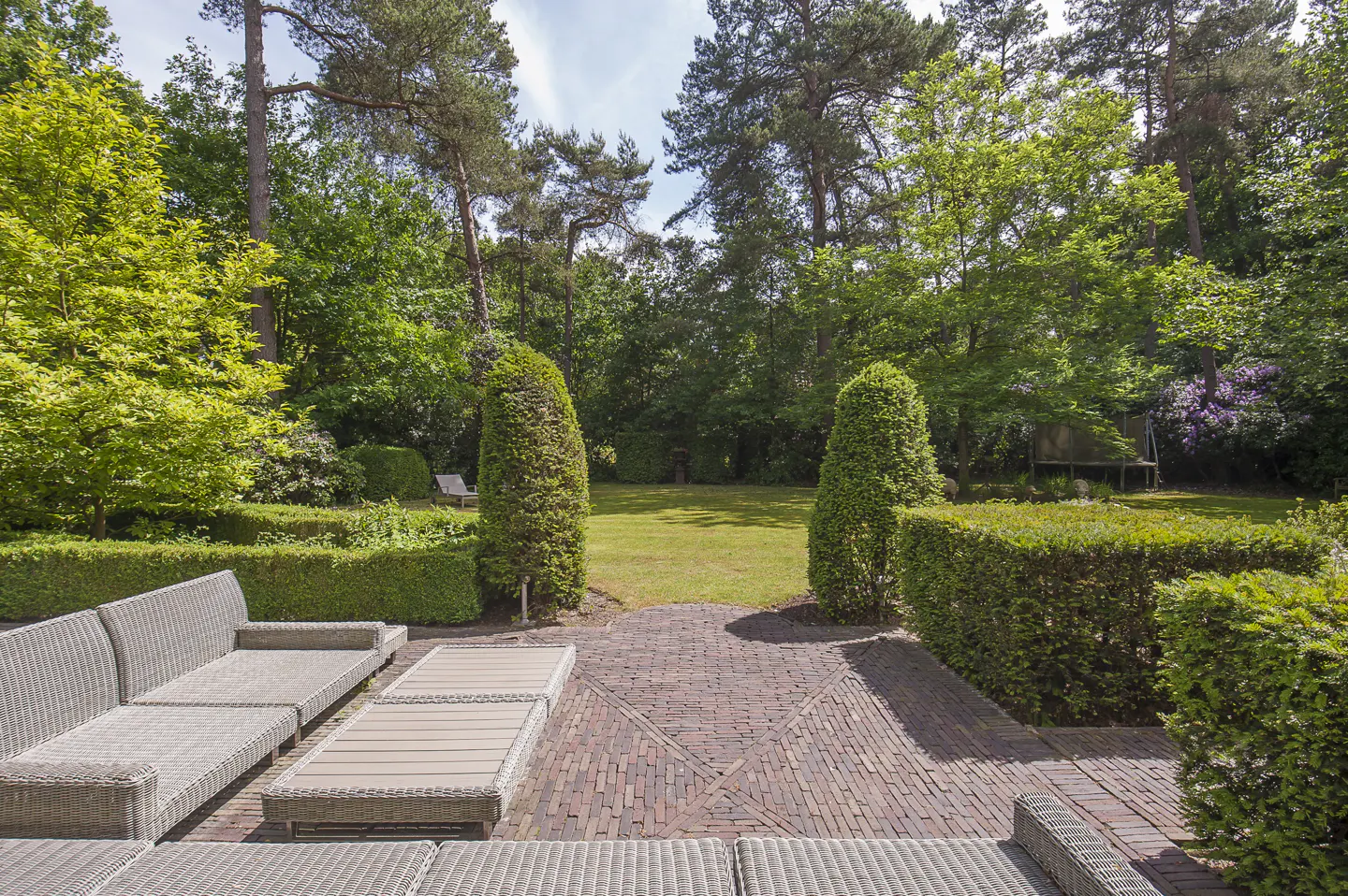 Outdoor patio with wicker furniture, brick pavers, and manicured hedges leading to a green lawn and trees.