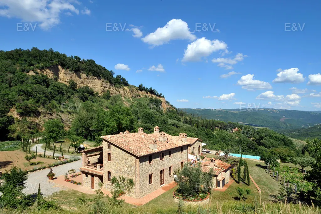 Stone house with a red tile roof, surrounded by green hills and a blue sky with white clouds. A pool is visible in the background.